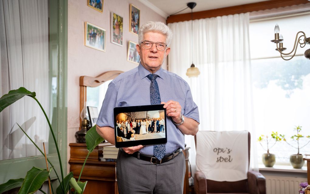 Man met een grijze kuif, een overhemd en een stropdas. Hij houdt een foto vast waarop negen stellen van hetzelfde gezin staan van wie hij het huwelijk sloot.
