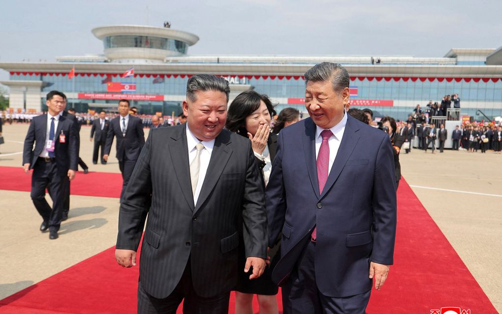 Twee mannen, de Noord-Koreaanse president en de Chinese president, wandelen glimlachend naast elkaar met achter hen een vertaalster. Op de achtergrond begeven zich nog veel andere personen en de terminal behorend tot Pyongyang International Airport.