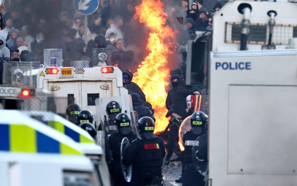 Gemaskerde personen, met vuur achter hen, staan tegenover de politie. Achter de gemaskerde personen bevinden zich nog meer demonstranten.