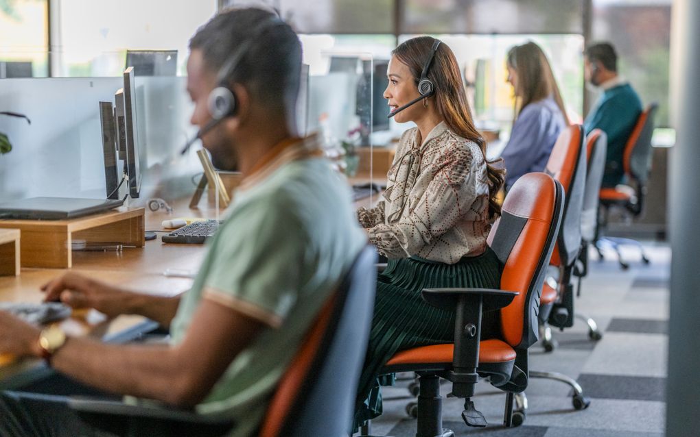 Mannen en vrouwen, met een headset op, werken in een modern callcenter en zitten op rode bureaustoelen.