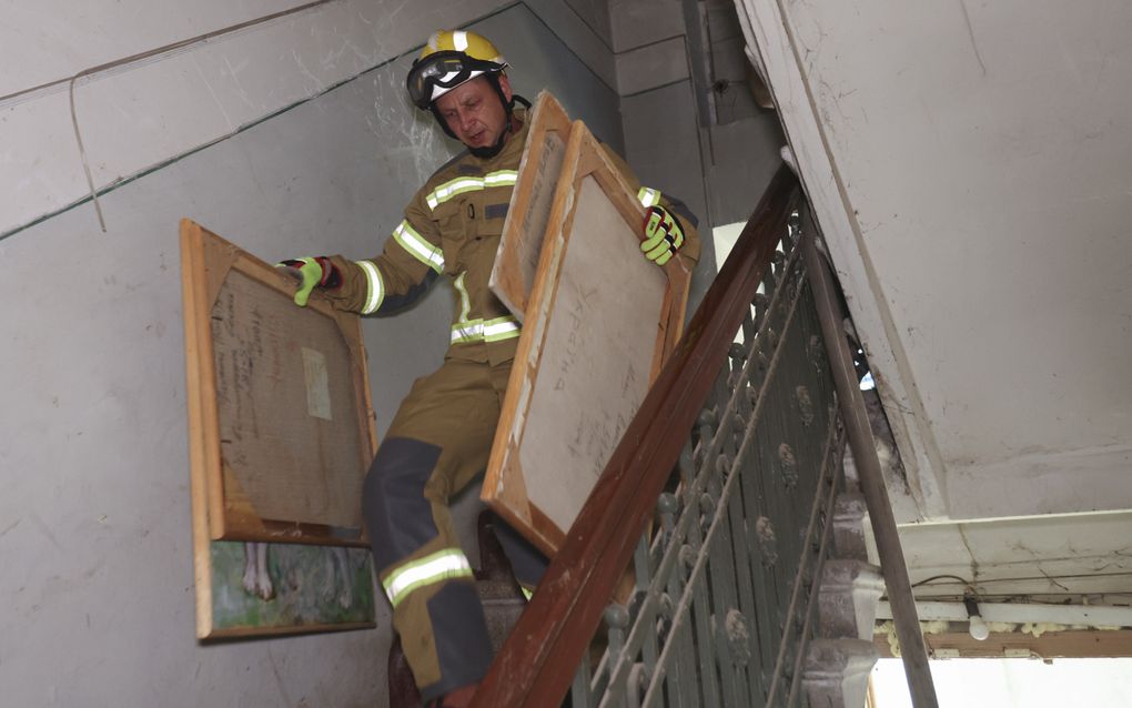 Een man met een helm op en een pak aan met reflecterende strepen draagt vier rechthoekige schilderijen. Hij loopt naar beneden in een trappenhuis.