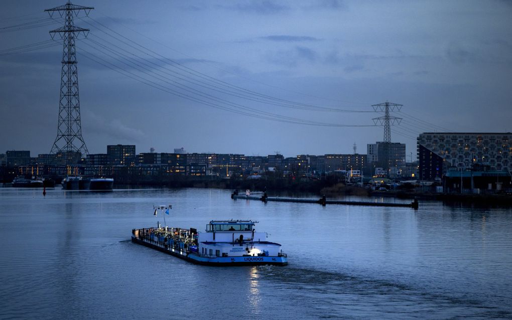 Een binnenvaartschip vaart in de schemering over een rivier. Op de achtergrond staan huizen en elektriciteitsmasten.