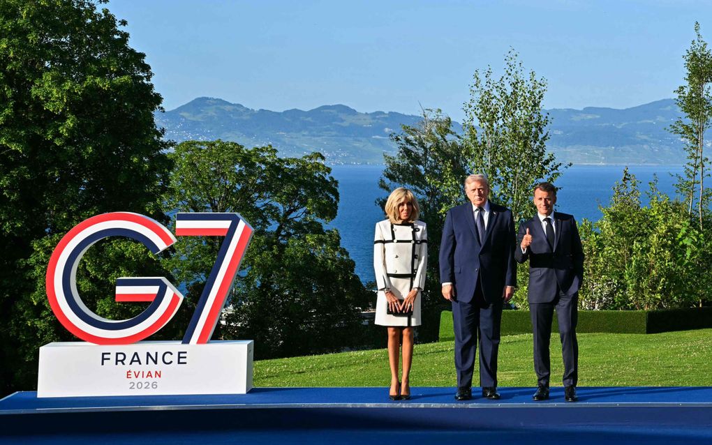 Twee mannen in een pak en een vrouw in een wit mantelpakje poseren bij een bord met de tekst G7 erop. Op de achtergrond zijn bergen en een meer zichtbaar.