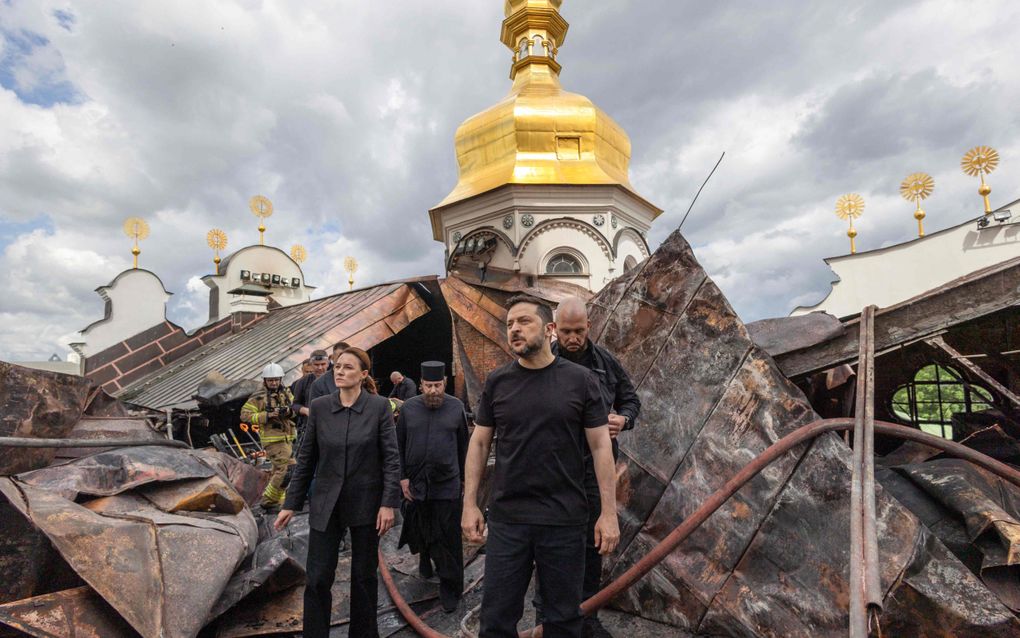 Mannen en vrouwen in het zwart, onder wie de Oekraïense president Zelensky, lopen tussen puin met op de achtergrond een toren met een gouden koepel.