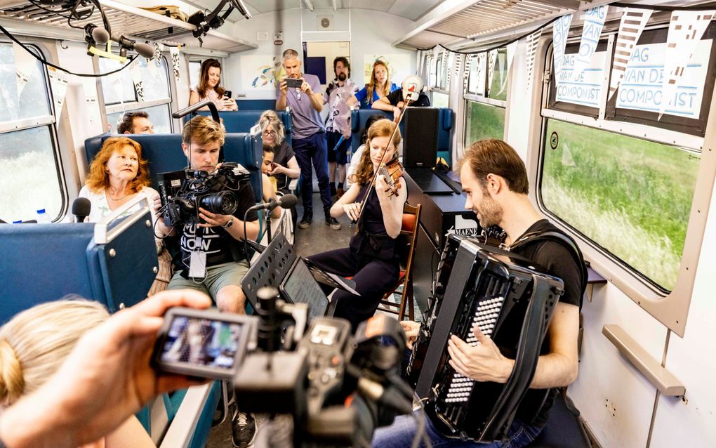 In een trein spelen twee musici respectievelijk accordeon en viool. Mensen luisteren. De musici worden gefilmd.