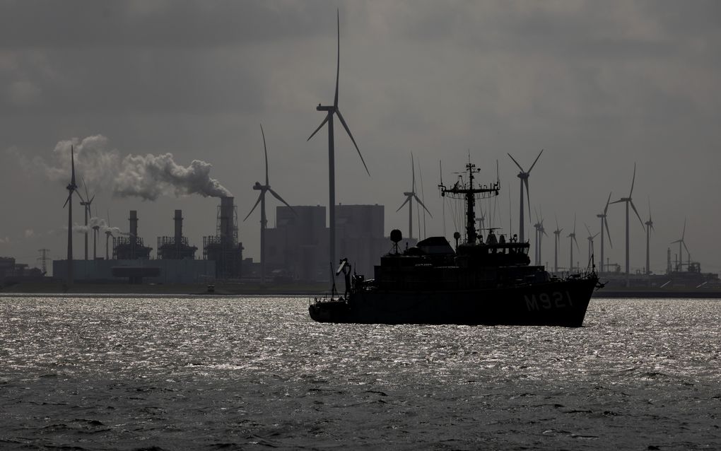 Een schip vaart in de avondschemering voor een industrieel havencomplex met rokende schoorstenen en windmolens op de achtergrond.