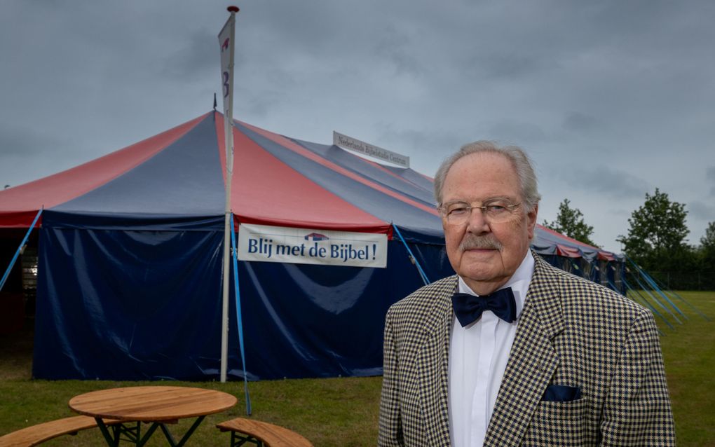 Een man met grijs haar, snor, geruit colbert, overhemd en vlinderstrik staat voor een grote rood-met-blauwe tent.