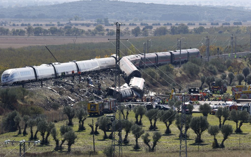 Twee treinen die op elkaar gebotst zijn. Eén staat nog half op de rails, de andere ligt er in stukken naast. Eromheen is veel ravage. Er lopen reddingswerkers en andere mensen.