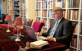 Man in grijs pak achter een laptop op een tafel met twee schemerlampen met rode kapjes. 