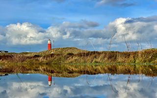 Rode vuurtoren van Texel met duinen op de voorgrond. De weerspiegeling is te zien in het water. 