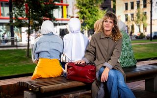 Vrouw met goudblond krulhaar, een bruine jas en een rode dokterstas zit op een houten bank langs een groenstrook. Achter haar op de bank zitten, met de rug naar haar toe, vier vrouwen met hoofddoeken. 