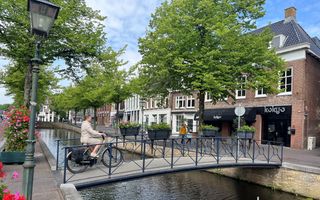Een vrouw fietst op een bruggetje over een gracht. Links op de kant staan bloembakken met rode en roze bloemen, aan de brug hangen ook rode en roze planten in een plantenbak.