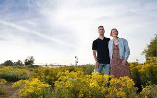 Een man en een vrouw staan in de uiterwaarden, tussen gele bloemen. In de verte een brug met twee bogen. 