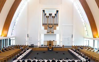 Interieur van modern, boogvormig kerkgebouw met in het midden vergadertafels met veertig mannen eromheen.