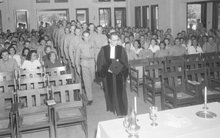 Predikant in toga loopt voor de militairen door het middenpad, terwijl kerkgangers toekijken.
