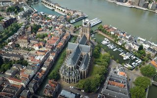 Luchtfoto van de binnenstad van Dordrecht met in het midden een groot kerkgebouw met daaromheen bomen, huizenrijen, omsloten door een gracht met een rivier die bovenlangs stroomt op het beeld.
