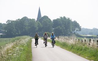 Drie fietsers rijden over een asfaltweggetje over een lage dijk. De foto is van voren genomen. Op de achtergrond zijn bomen te zien met een kerktoren die erbovenuit steekt. De zon staat hoog aan de hemel.
