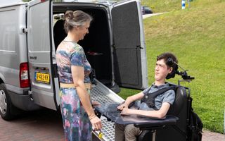 Moeder en zoon staan voor een grijze bus. De jongen zit in een elektrische rolstoel.