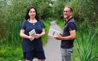 Een vrouw met halflang donker haar en in het donkerblauw gekleed staat op een pad tussen groene bomen. Ze heeft een boek in haar hand. Rechts staat een man met bril en een blauw poloshirt. Hij kijkt schuin opzij en heeft ook een boek in zijn handen. 