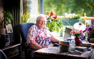 Een oude vrouw met rood-wit-blauwe kleding zit op een bureaustoel aan een tafel naast het raam. De tafel staat vol met boeken, bakjes en bloemen.