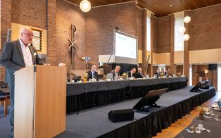 Een man in grijs pak staat achter een katheder; daarnaast een lange tafel waarachter predikanten zitten.