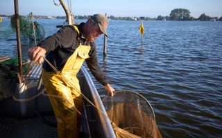 Ale de Jager, man met petje en gele waterdichte werkkleding aan, staat in zijn boot op een meer. Hij houdt een visnet boven water.