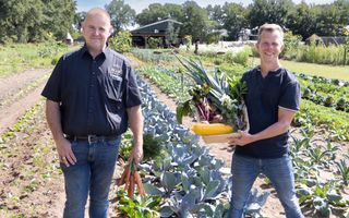 Twee mannen staan met groenten in hun handen in een groentetuin. Op de achtergrond bloeiende bloemen en een veranda.