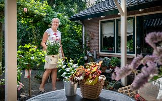 Lachende vrouw met wit shirt en lichte rok tot op de knie houdt een pot met bloemen vast. Ze staat in een tuin met veel struiken en bloemen.