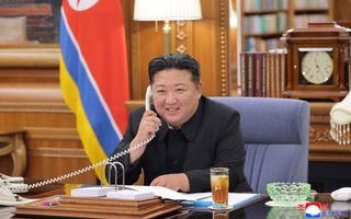 Een man in pak zit lachend achter een bureau aan de telefoon. 