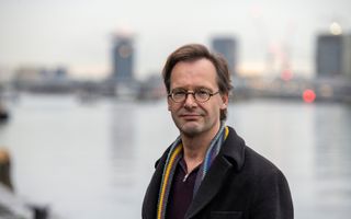 Een man met bruin haar en een bril kijkt enigszins peinzend in de lens, op de achtergrond (vaag weergegeven) een wateroppervlak.