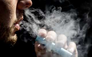 Een bebaarde man houdt een vape vast, een flinke rookwolk komt uit zijn mond.