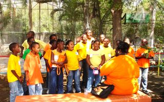 Een groep scholieren is gekleed in oranje en gele shirts. Ze luisteren naar een docent op een bankje voor hen. De docent draagt een oranje T-shirt. 