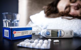 Een doosje paracetamol, een koortsthermometer en een glas water staan op een nachtkastje. Op de achtergrond ligt een vrouw op bed.