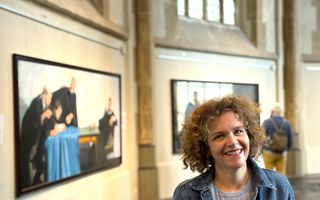 Lachende vrouw, krullend haar, spijkerjasje. Op de achtergrond twee van de acht schilderijen met de werken van barmhartigheid.