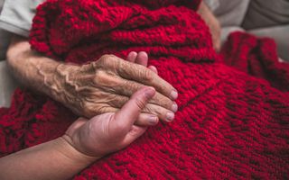 Een kwetsbare oude man met een gebreid rood kleed over zich heen houdt de hand van een vrouw vast.