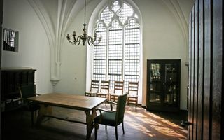 Ruimte met een hoog gotisch raam, houten vloer en kroonluchter die in het midden boven een houten tafel hangt met aan twee kanten houten stoelen met groene bekleding.