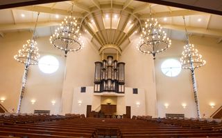 Een kerkzaal met lege, bruine kerkbanken, een bruine kansel, een orgel en kroonluchters.