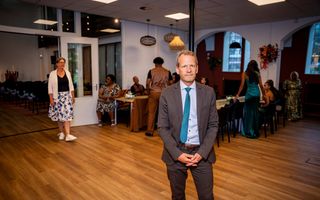 Man in donker pak met stropdas staat in een ruimte met op de achtergrond mensen rond een tafel.