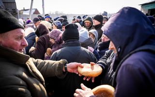 Groep mensen staat dicht bij elkaar, dik aangekleed en met mutsen op. Er worden broden uitgedeeld.