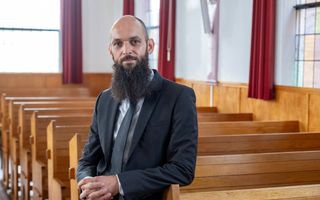 Dominee Schakel, een man met zwarte baard, gekleed in donker kostuum, leunt tegen een kerkbank.