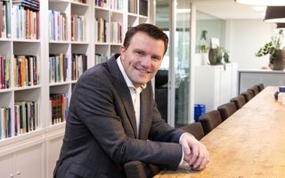 Een man met een donker pak en een wit overhemd kijkt glimlachend naar de camera. Hij leunt op een stoel achter een houten tafel. 