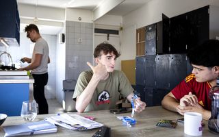 Vier jongemannen in een rommelige kamer. Twee zitten er aan tafel, twee staan achterin bij het aanrecht. Op tafel liggen papieren en andere spullen.