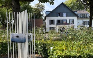 Borden aan witte spijlen. Achter de struiken een wit gebouw.