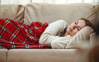 Vrouw ligt met gesloten ogen op een beige bank. Ze heeft een rood-zwart-wit geblokte plaid over zich heen getrokken.