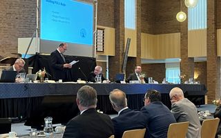 Vier mannen achter een tafel luisteren naar dominee Buijs, die staand achter een lange tafel en met een A4’tje in de hand een vergadering afsluit.