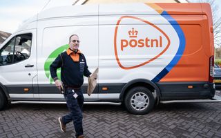 Een medewerker van PostNL loopt met een pakketje over een klinkerstraat. Zijn bus met het logo van PostNL staat achter hem. 