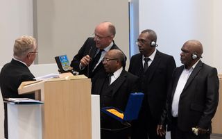 Vijf mannen in zwarte pakken bekijken een present in de vorm van een gedenkplaatje. Drie mannen zijn zwart, twee wit.