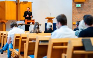 Dominee Hoogerbrug, een man in zwart kostuum, staat achter een tafel voorin een kerk, terwijl mensen op kerkbanken naar hem luisteren. 