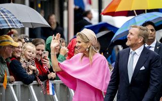 De koning in blauw pak, de koningin in roze outfit. Links publiek met paraplu's.