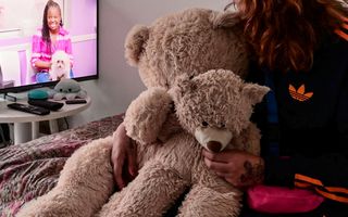 Een jonge vrouw zit met twee beren in haar hand op bed en kijkt naar een tv-programma.